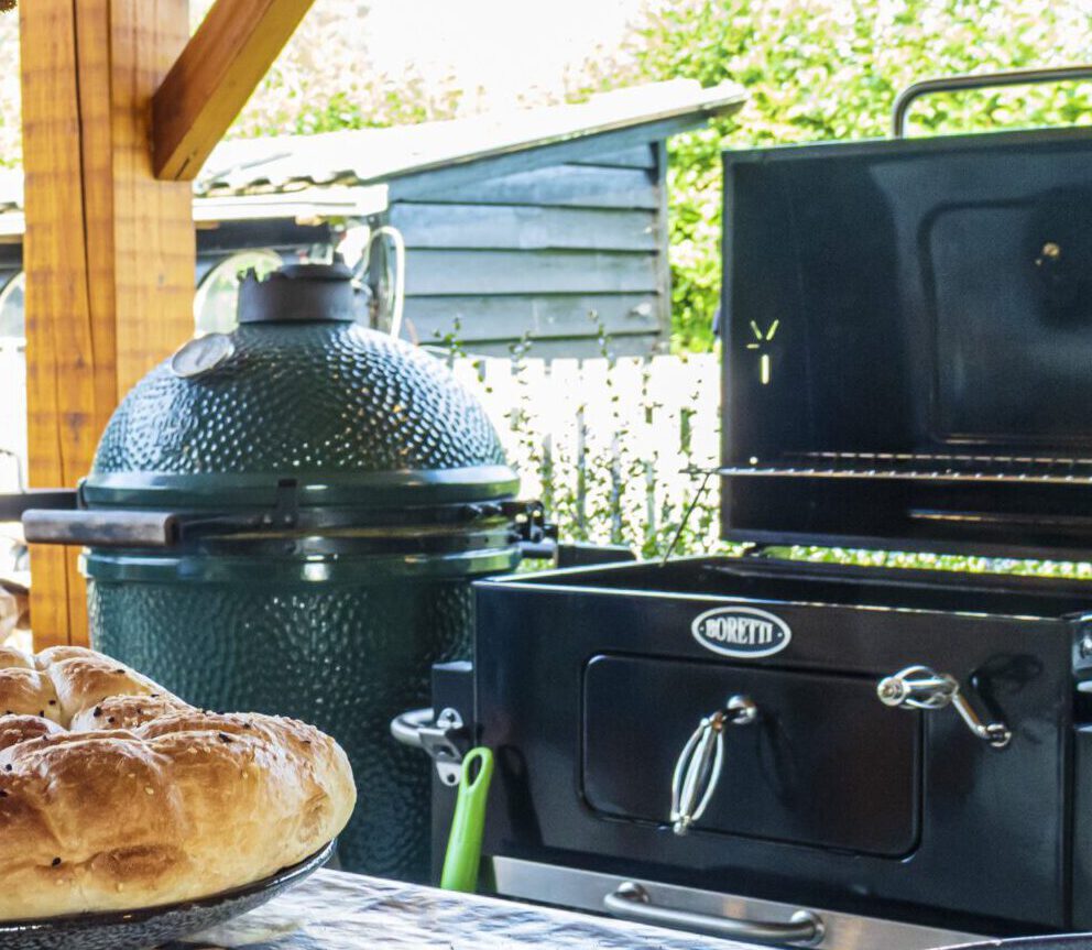 BBQ-setup met kamado, oven en brood op tafel onder overkapping.