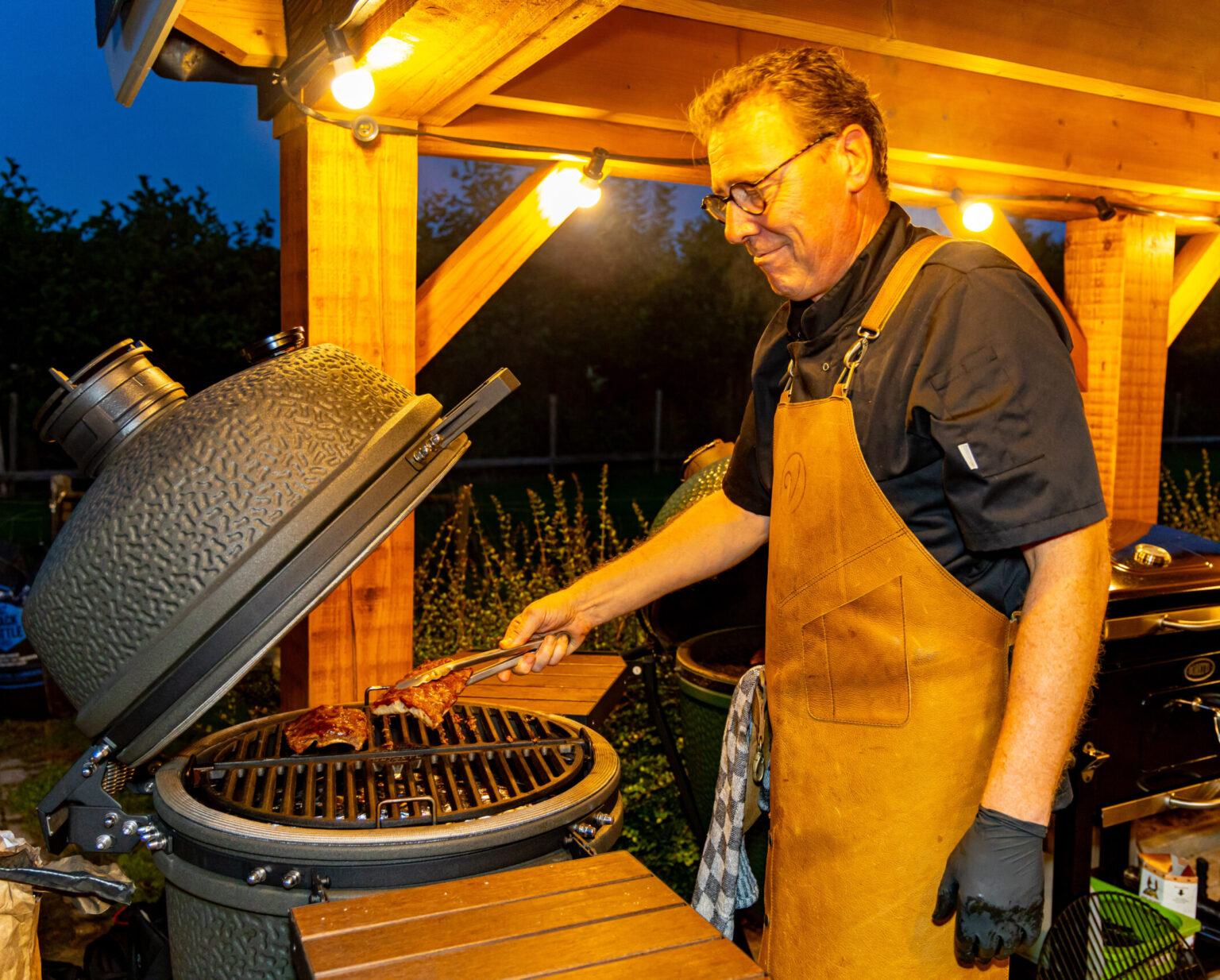 Man grillt vlees op een grote kamado barbecue buiten in de avond.